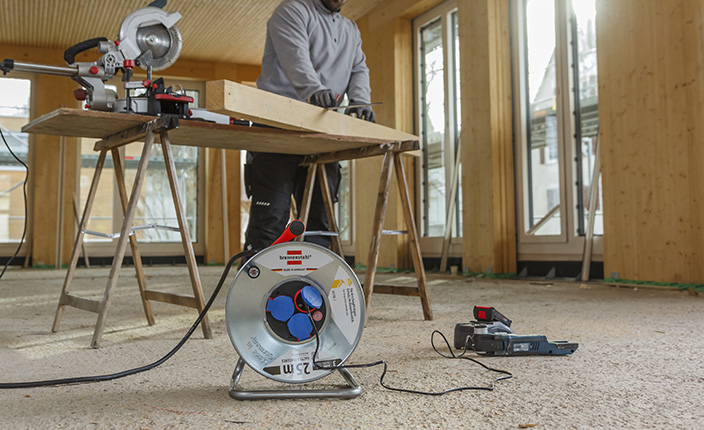 Ein Mann arbeitet an Holz in einem Raum und im vordergrund sieht man eine Kabeltrommel