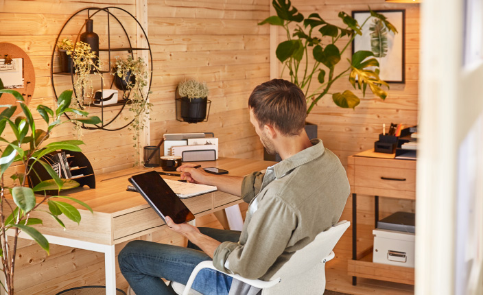 Ein Mann arbeitet im Gartenhaus am Schreibtisch und nutzt dabei Tablet und Notizbuch. Der Arbeitsplatz ist mit Pflanzen, Deko und Holzregalen gemütlich eingerichtet.