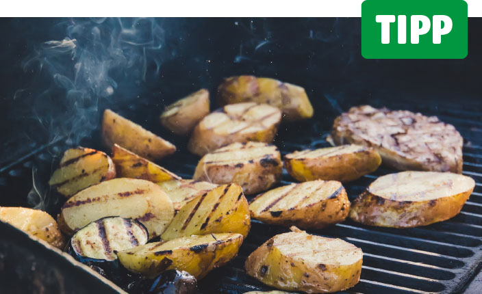 Mehrere halbierte Kartoffeln mit Grillstreifen liegen auf einem Grillrost, daneben ein Stück Fleisch. Zwischen den Kartoffeln steigt Rauch auf. Oben rechts steht in weißer Schrift auf grünem Grund: Tipp