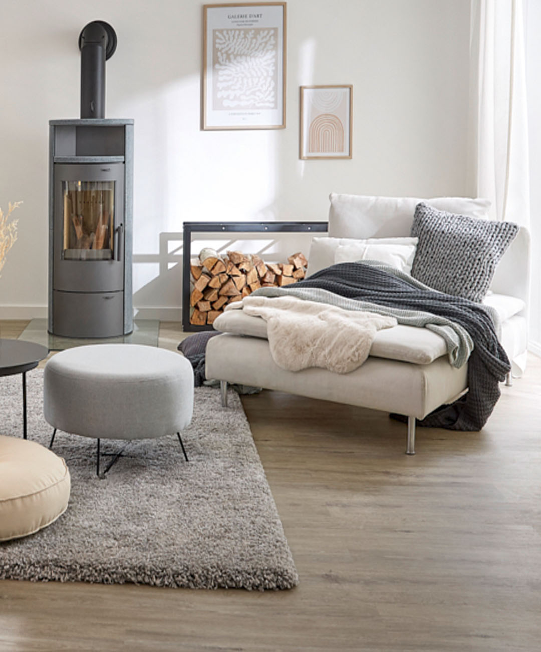 Wohnzimmer mit weißer Wand, hellbraunem Laminatboden, Kamin, Bildern, Feuerholz-Aufbewahrung, grauem Teppich und weißem Liegesofa; In der oberen rechten Ecke des Bildes die Aufschrift auf weißen Hintergrund „Innenbereich“