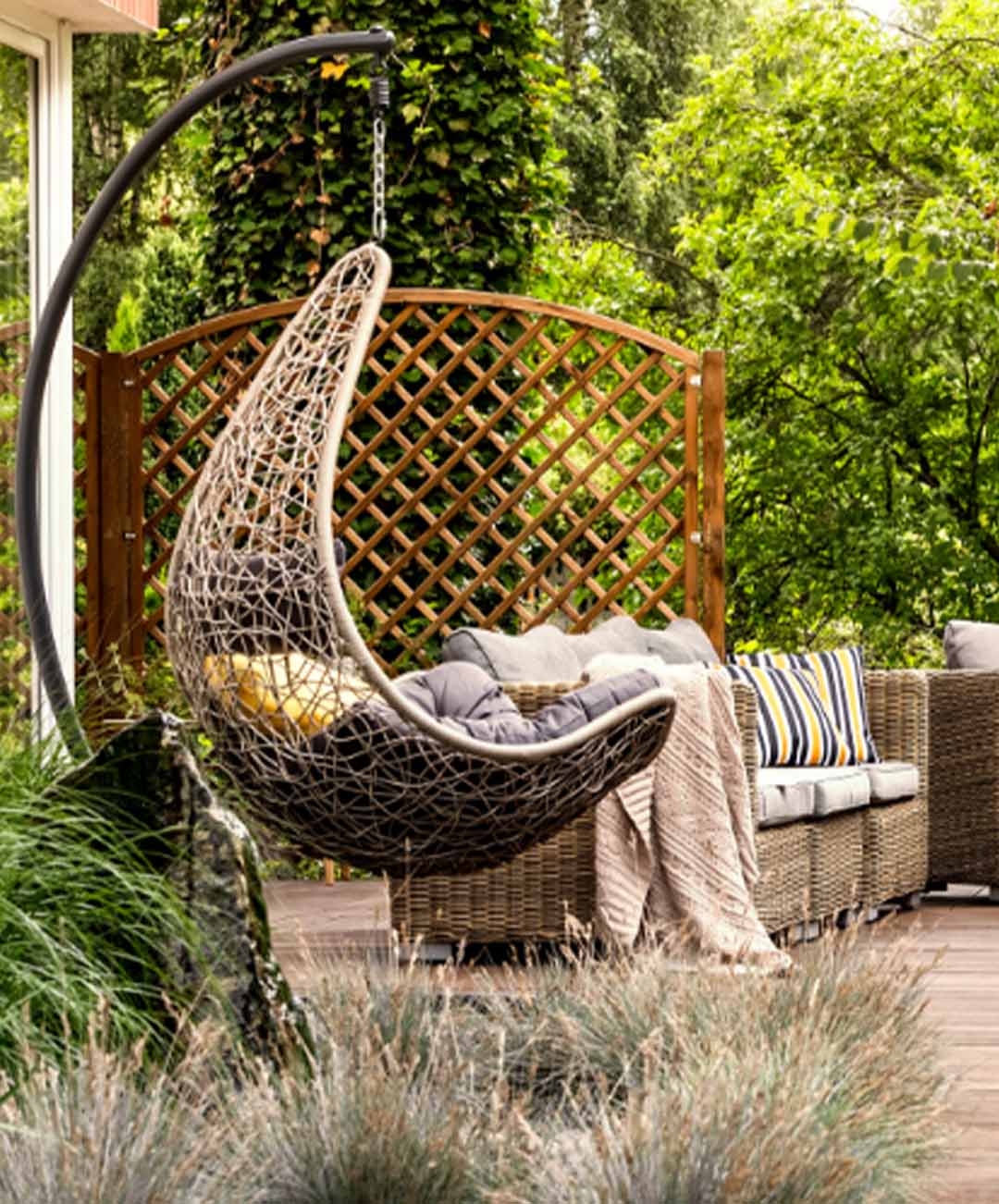 Ein Terrassenausschnitt mit kunstvoll geflochtenen Hängesessel, daneben ein geflochtenes Sofa mit Sitzkissen; verschiedenen Pflanzenarten im Hintergrund; In der oberen rechten Ecke des Bildes die Aufschrift auf weißen Hintergrund „Aussenbereich“