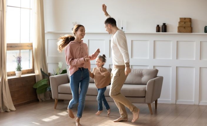 Vater, Mutter und Tochter tanzen in einem gut beleuchtetem Wohnzimmer