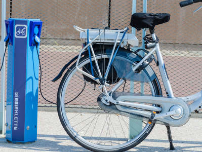 Ein silbernes E-Bike ist an einer blauen Ladestation angeschlossen. Die Station steht auf einem gepflasterten Untergrund vor einem Zaun, der Akku des E-Bikes befindet sich über dem Hinterrad