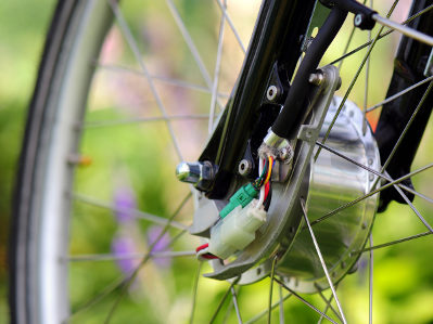 Ein E-Bike-Vorderrad mit einem eingebauten Nabenmotor ist aus der Nähe zu sehen. Die elektrischen Anschlüsse und Kabel sind gut sichtbar, im Hintergrund ist unscharf grüne Natur zu erkennen
