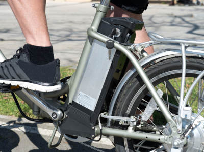 Ein E-Bike mit silbernem Rahmen wird aus der Seitenansicht gezeigt. Im Fokus steht der große Akku hinter dem Sattelrohr. Ein Fahrer mit schwarzen Sportschuhen sitzt auf dem Fahrrad