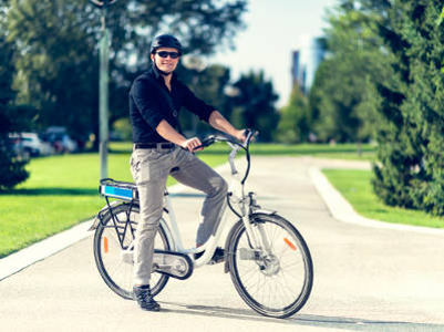 Ein Mann mit schwarzem Hemd, grauer Hose und Helm sitzt auf einem silbernen E-Bike in einem Park. Er lächelt und trägt eine Sonnenbrille, im Hintergrund sind Bäume und Hochhäuser zu sehen