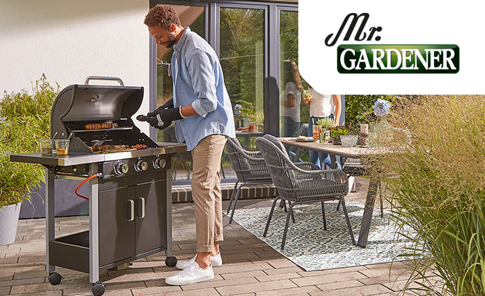 Ein Mann steht an einem Grill und grill in einem Garten hintergrund sieht man eine  und einen Gartentisch mit Stühlen