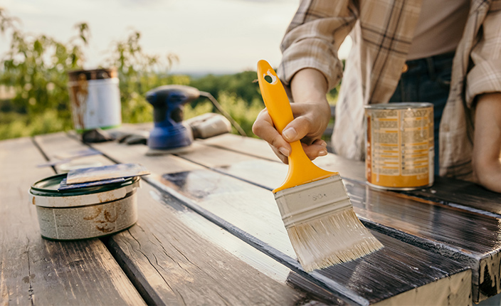 Eine Person streicht eine Holzfläche mit einem breiten Pinsel und transparenter Lasur – auf dem Tisch stehen Schleifgerät, Dosen und Schleifpapier, im Hintergrund ist grüne Landschaft zu sehen.