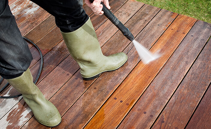 Eine Person in Gummistiefeln reinigt eine Holzterrasse mit einem Hochdruckreiniger – der Wasserstrahl lässt den Unterschied zwischen gereinigter und verschmutzter Fläche deutlich erkennen.