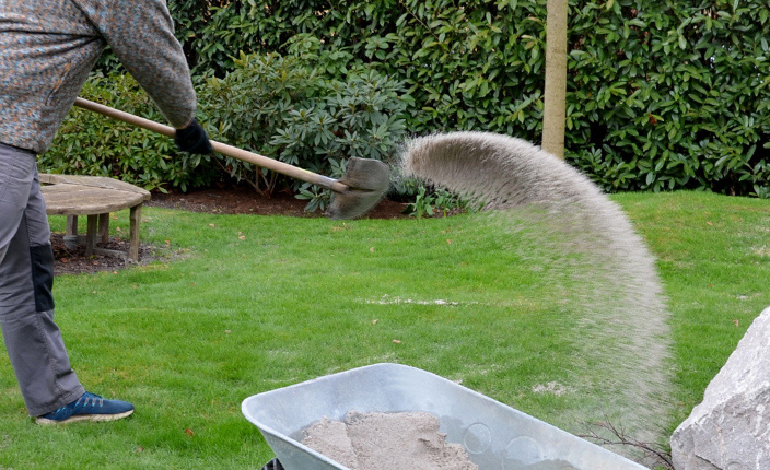 Ein Person schüttet mit einer Schaufel Sand auf einen Rasen