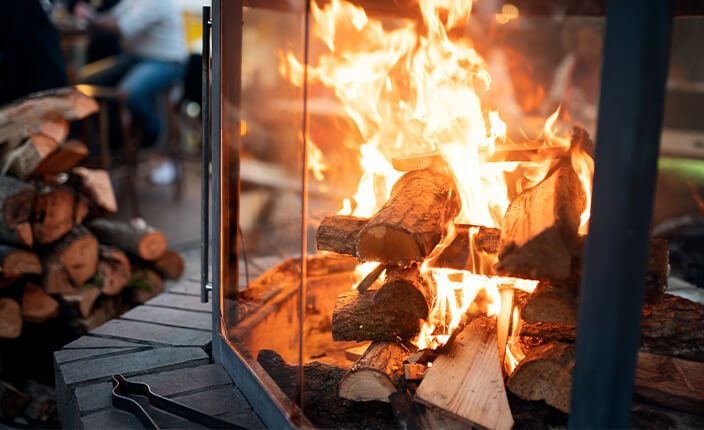 Loderndes Kaminfeuer in einem verglasten Außenkamin mit gestapeltem Brennholz im Hintergrund.