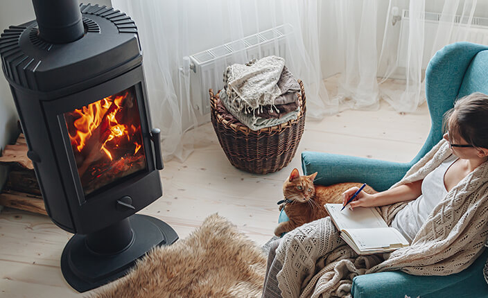 Gemütliche Szene mit brennendem Kaminofen, Frau auf einem Sessel beim Schreiben, eingehüllt in eine Decke, mit einer Katze auf dem Schoß.