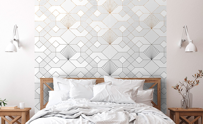 Schlafzimmer mit hellem Holzbett, weißen Kissen und Decken. Im Hintergrund eine großflächige Tapete mit geometrischem Muster in Gold- und Silbertönen. Links und rechts des Betts sind weiße Wandleuchten sowie Nachttische mit Pflanzen und Deko zu sehen