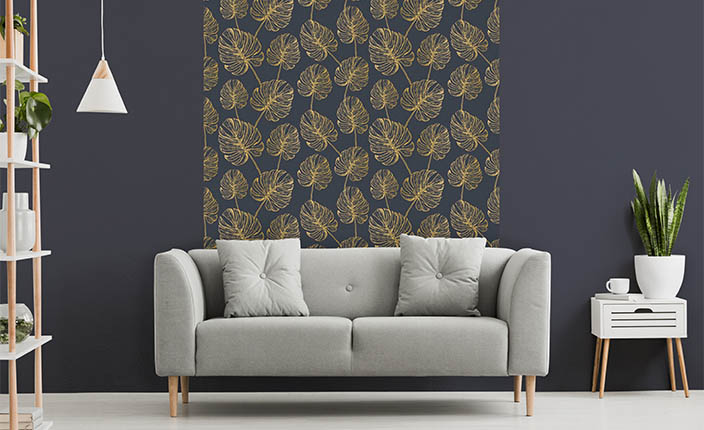 Modernes Wohnzimmer mit dunkelblauer Wandfarbe und einer Akzenttapete mit goldfarbenem Blattmuster hinter einem hellgrauen Sofa. Rechts steht ein weißer Beistelltisch mit Pflanze und Tasse, links ein weißes Regal mit Dekor.