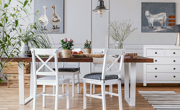 Heller Essbereich im Landhausstil mit einem rechteckigen Holztisch und weißen Stühlen mit schwarzen Sitzkissen. Auf dem Tisch stehen Körbe, Blumenvasen und Obst. Links und rechts an der Wand hängen Tiergemälde, darunter weiße Kommoden. Eine schwarze Pendelleuchte hängt zentral über dem Tisch. Der Raum ist mit Pflanzen und Holzboden wohnlich gestaltet