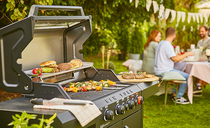 Ein Grill im Vordergrund mit essen drauf und einigen Personen im Hintergrund