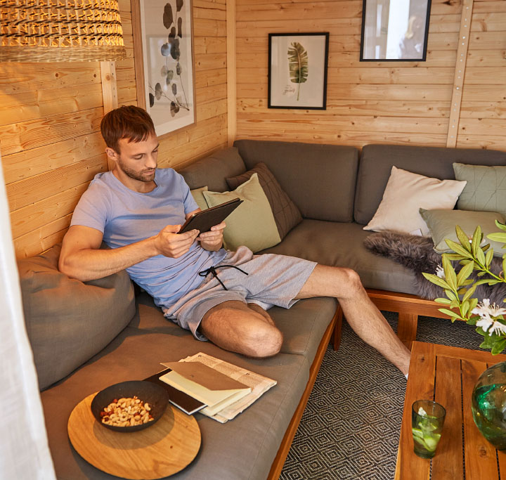Ein Mann sitzt entspannt in einem modern eingerichteten Gartenhaus auf einem grauen Ecksofa und liest auf einem Tablet. Auf dem Couchtisch stehen Getränke, ein Snack und Notizbücher. Im Hintergrund sind dekorative Pflanzen und gerahmte Naturmotive an der Holzvertäfelung zu sehen.