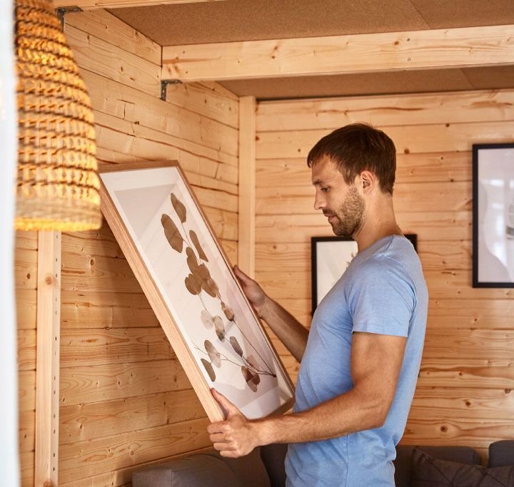 Ein Mann in hellblauem T-Shirt hängt in einem Holz-Gartenhaus ein gerahmtes Bild mit botanischem Motiv an die Wand.