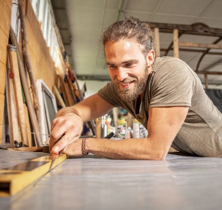 Ein junger Mann beim Heimwerken, der lächelnd mit einem Cuttermesser und einer Wasserwaage Bodenmaterial für ein Gartenhaus zuschneidet.