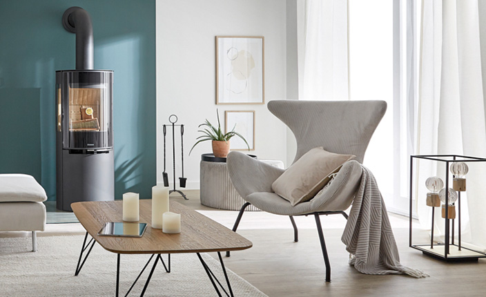 Gemütliches Wohnzimmer im hellen Einrichtungsstil; Ein Kamin an dunkler, türkiser Wand, ein Sessel neben einem Cordhocker
