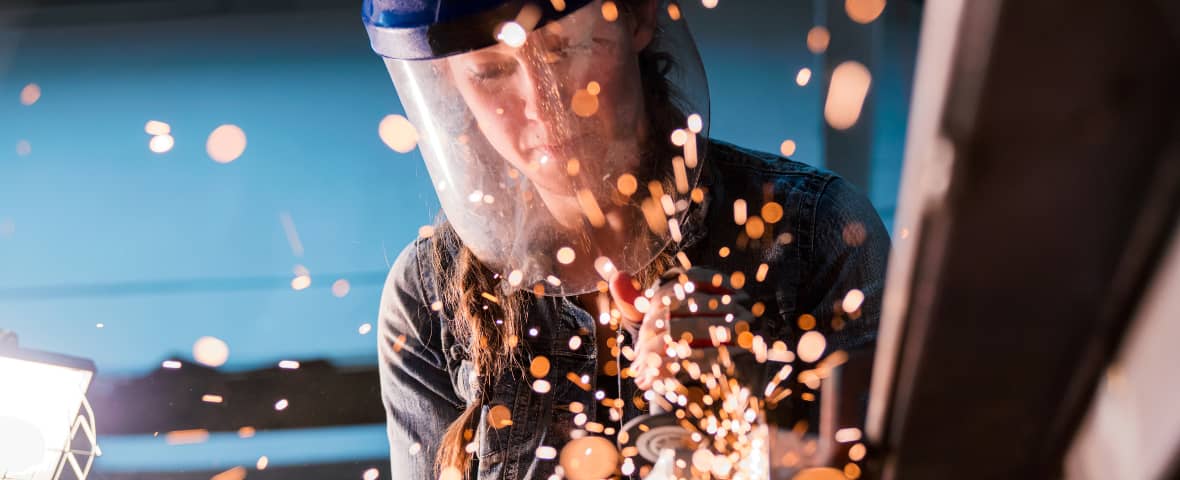 Eine Frau mit Schutzvisier schweißt an einem Metallteil. Um sie herum fliegen helle Funken, die Szene ist in kräftigem Licht und vor blauem Hintergrund aufgenommen