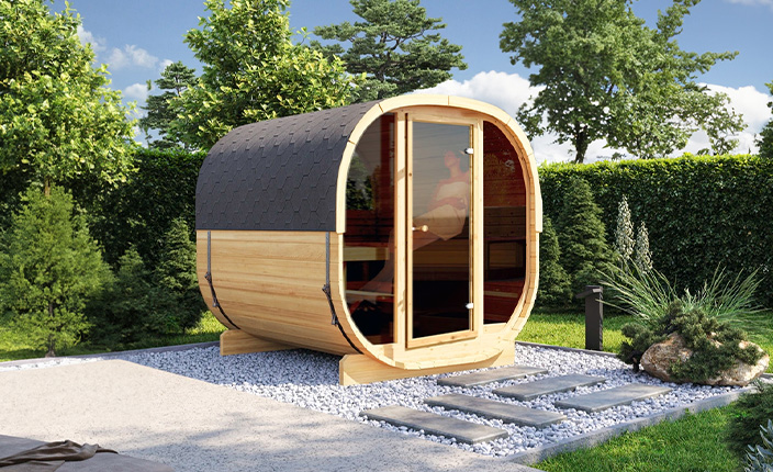 Moderne Fasssauna aus Holz im Garten auf Kiesfläche, umgeben von Hecken und Bäumen, mit Trittsteinen und gepflegtem Grün.