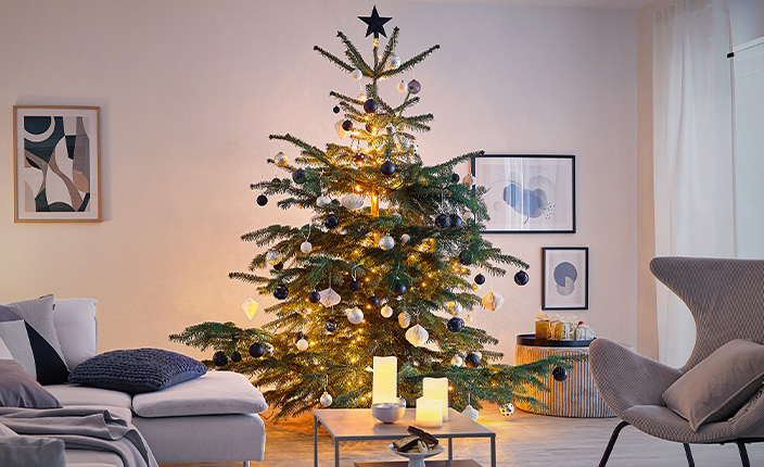 Ein weihnachtlich geschmückter Tannenbaum steht in einem modern eingerichteten Wohnzimmer. Der Baum ist mit warmweißer Lichterkette, schwarzen und silbernen Kugeln sowie weißen Anhängern dekoriert. Um ihn herum stehen Kerzen und ein kleiner Geschenkkorb. Im Hintergrund hängen gerahmte Bilder an einer hellen Wand