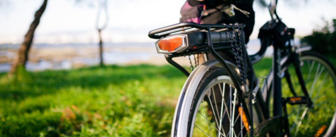 Ein E-Bike steht auf einer Wiese in der Nähe eines Gewässers. Im Vordergrund ist der Gepäckträger mit integriertem Akku und Rücklicht zu sehen, im Hintergrund verschwimmt die Landschaft leicht
