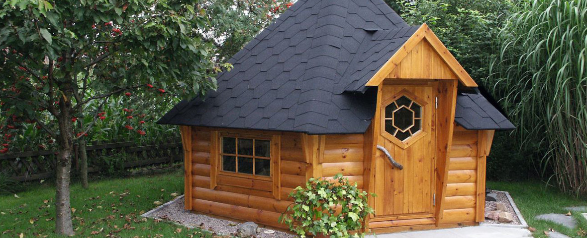 Eine sechseckige Grillkota aus naturbelassenem Holz mit spitzem Dach und zentralem Schornstein steht auf einer Rasenfläche. Die Tür mit kleinem Fenster ist mittig platziert, flankiert von zwei weiteren Fenstern.