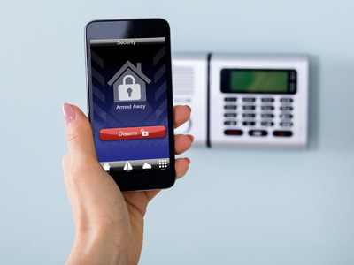 Eine Hand hält ein Smartphone mit aktivierter Sicherheits-App vor einer modernen Alarmanlage an der Wand