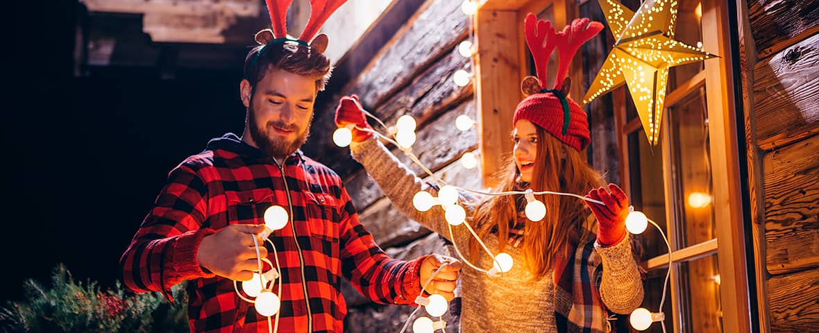 Ein Paar mit Rentier-Haarreifen dekoriert gemeinsam eine Holzhütte mit einer leuchtenden Lichterkette. Beide lachen, im Fenster hängt ein beleuchteter Papierstern