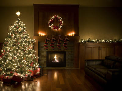 Ein geschmückter Weihnachtsbaum mit Lichterkette steht neben einem Kamin mit brennendem Feuer. Über dem Kamin hängen weihnachtliche Strümpfe, auf dem Sims stehen Kerzen, darüber ein beleuchteter Kranz. Lichterketten schmücken auch die Anrichte im Hintergrund