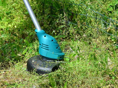 Eine elektrische Motorsense mit blauem Gehäuse kürzt hohes, trockenes Unkraut entlang eines Drahtzauns. Der Schneidkopf ist teilweise von Gras umgeben, der Einsatz erfolgt auf unebenem Gelände