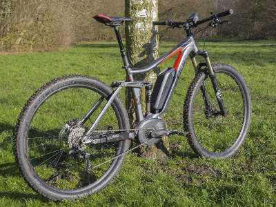 Ein sportliches E-Mountainbike mit Mittelmotor steht auf einer Wiese vor einem Baum. Der Rahmen ist grau mit roten Akzenten und die Reifen sind leicht verschmutzt vom Fahren im Gelände