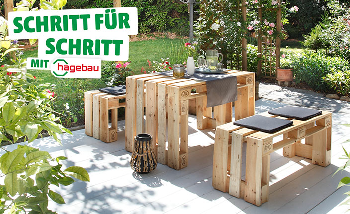 Gartensitzgruppe aus Europaletten bestehend aus einem Tisch und zwei Bänken mit schwarzen Sitzkissen, auf einer hellen Terrasse im Garten. Text: "Schritt für Schritt mit hagebau".