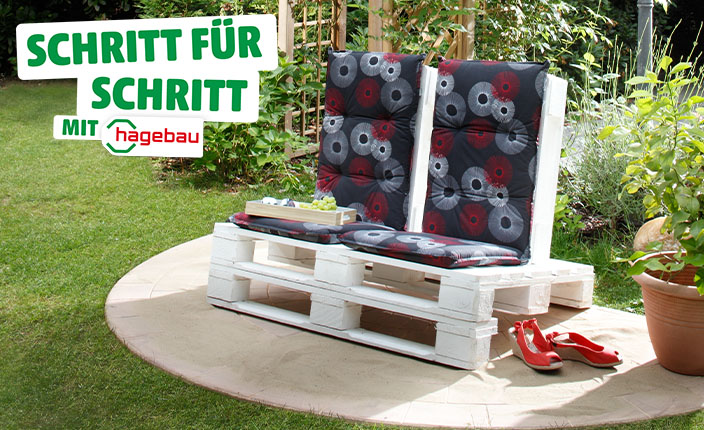 Selbstgebaute Gartenbank aus weißen Paletten mit schwarz-gemusterten Polstern, auf einer runden Terrasse im Garten. Daneben stehen rote Schuhe. Text: "Schritt für Schritt mit hagebau".