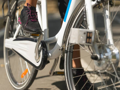 Ein weißes E-Bike wird im unteren Bereich gezeigt, während eine Person mit schwarzen Sportschuhen auf den Pedalen steht. Deutlich sichtbar sind der geschlossene Kettenkasten und Reflektoren an Pedalen und Reifen